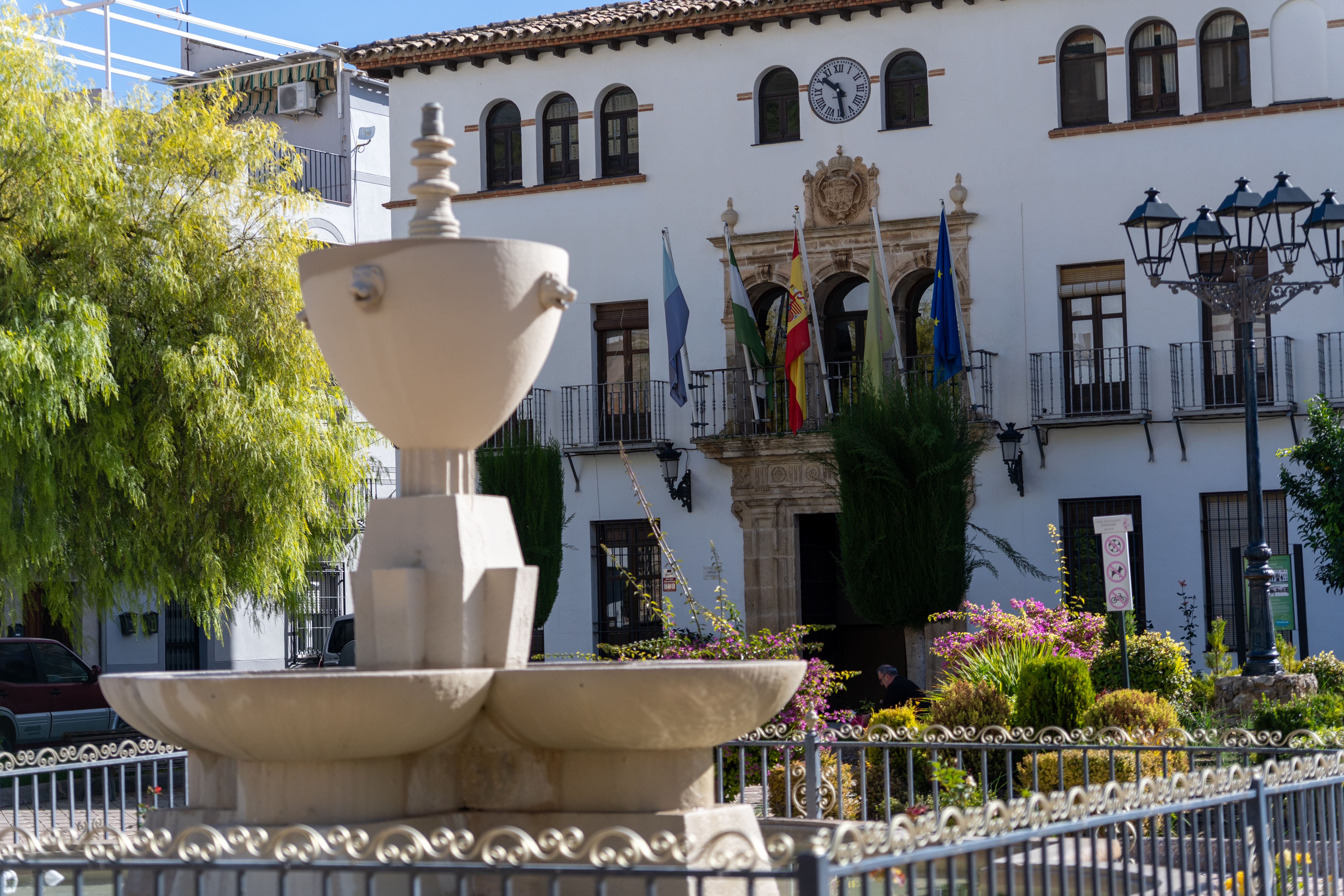 Fuente ornamental en primer plano, situada en una plaza ajardinada frente a un edificio institucional de fachada blanca con balcón, reloj y banderas. La escena muestra un espacio urbano cuidado, con flores, árboles y farolas tradicionales.