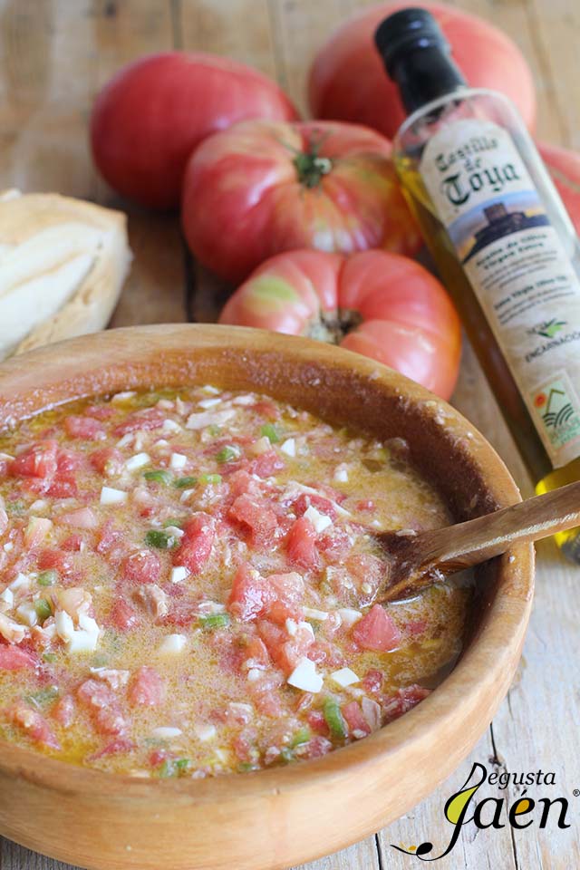 Pipirrana servida en un cuenco de barro, elaborada con tomate troceado, huevo, pimiento y aceite de oliva virgen extra. La imagen incluye una cuchara de madera y, al fondo, tomates frescos y una botella de aceite, destacando el carácter fresco y tradicional de esta ensalada de verano.