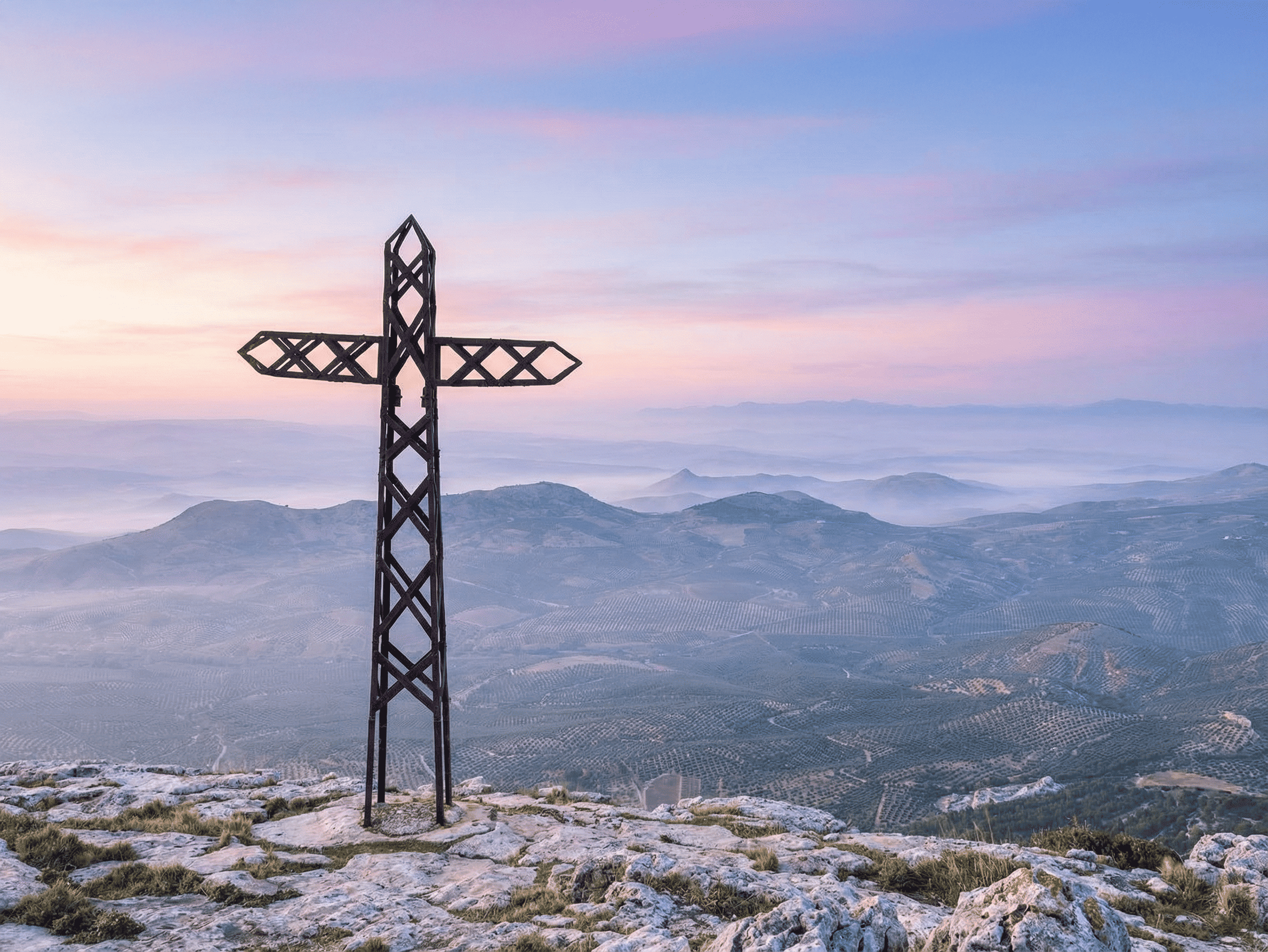 Una toma de la Cruz de la Atalaya en Jimena, Jaén, al amanecer. La estructura de hierro calado destaca sobre un paisaje de montañas neblinosas y el extenso mar de olivos de Sierra Mágina.