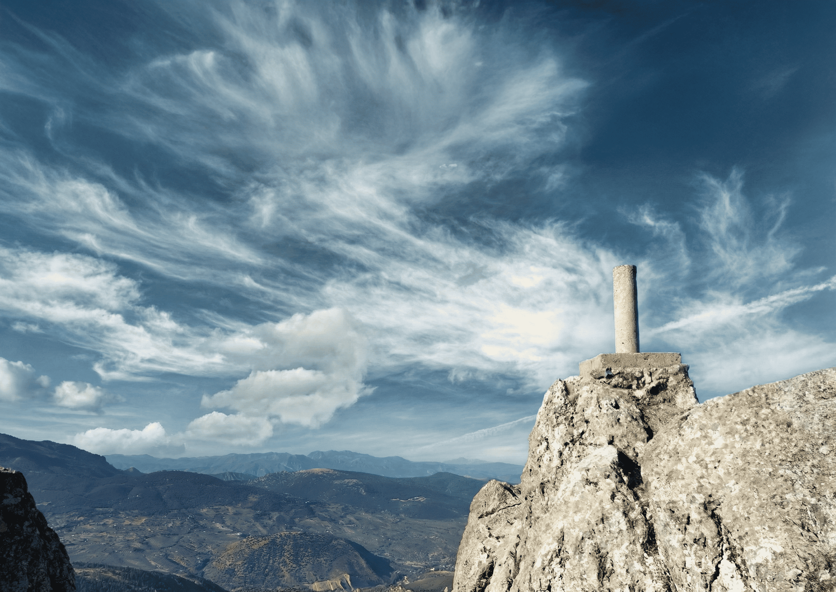 Vista panorámica desde una zona rocosa, con un vértice geodésico situado sobre una peña en primer plano. Al fondo se extiende un paisaje de sierras y valles bajo un cielo amplio cubierto de nubes blancas alargadas.