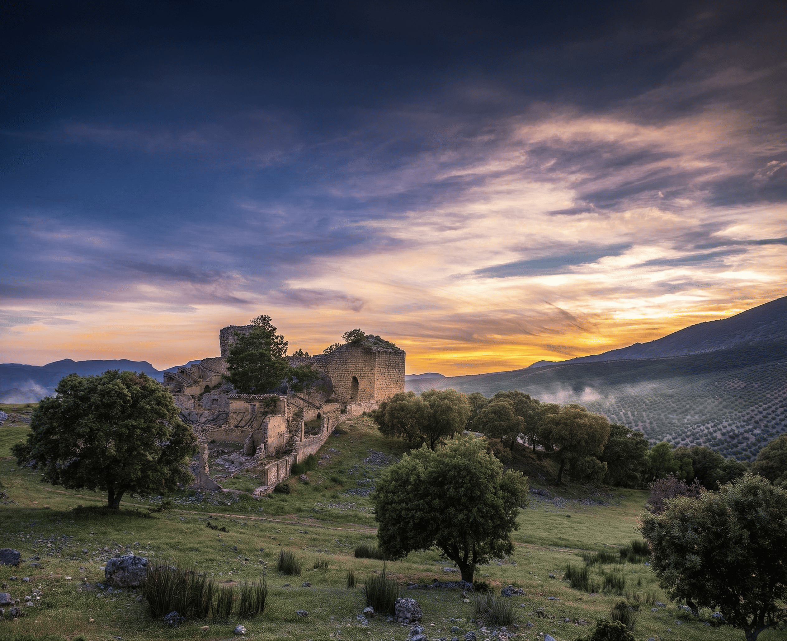 Ruinas de una construcción antigua de piedra situadas en un paisaje abierto con árboles dispersos y suaves lomas. Al fondo se aprecia un valle entre montañas bajo un cielo de atardecer con tonos azules, violetas y anaranjados.