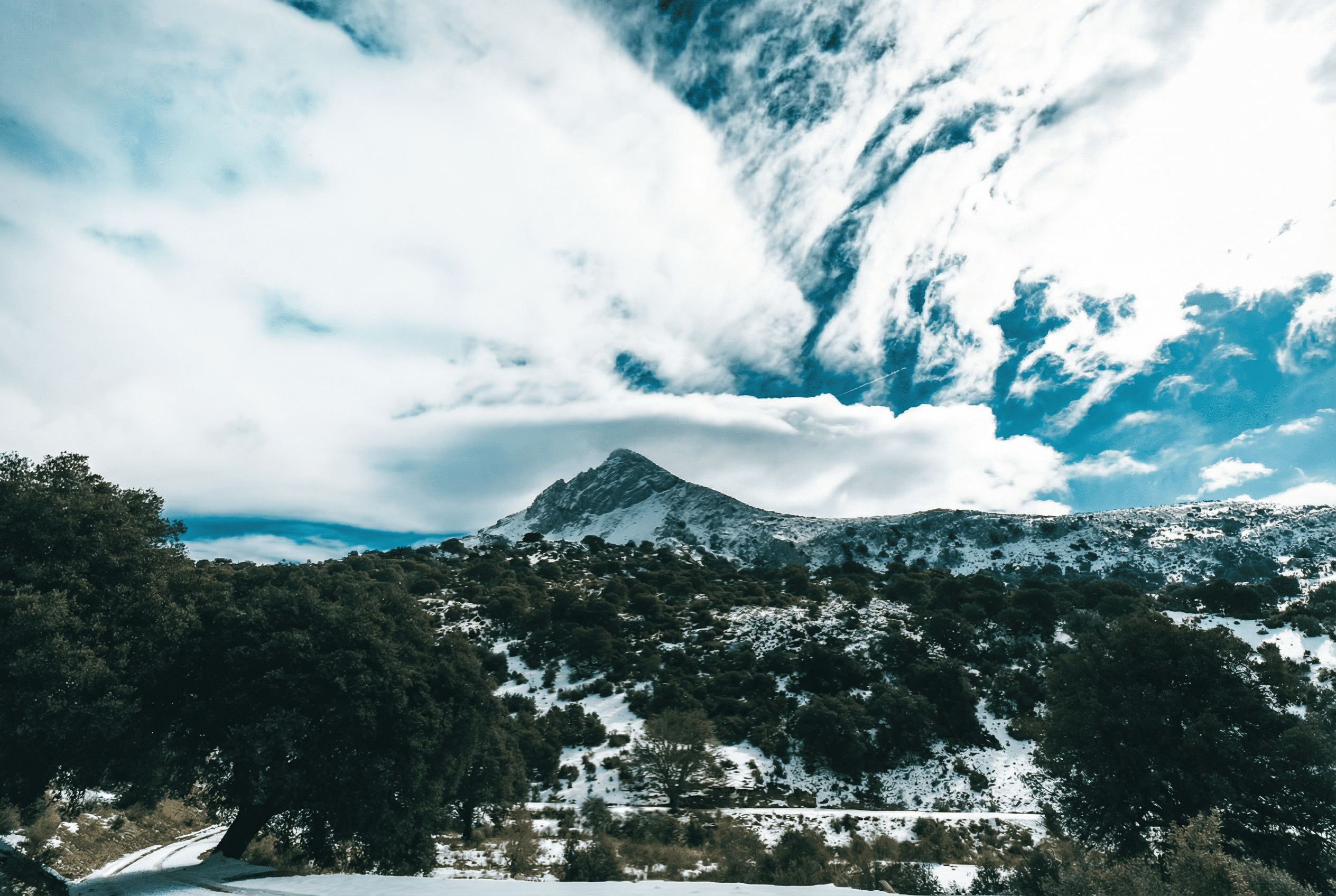 Paisaje de montaña con una cumbre nevada en el centro, rodeada de vegetación y zonas cubiertas de nieve. Sobre la escena destaca un cielo amplio con nubes blancas muy marcadas que contrastan con el azul intenso del fondo.