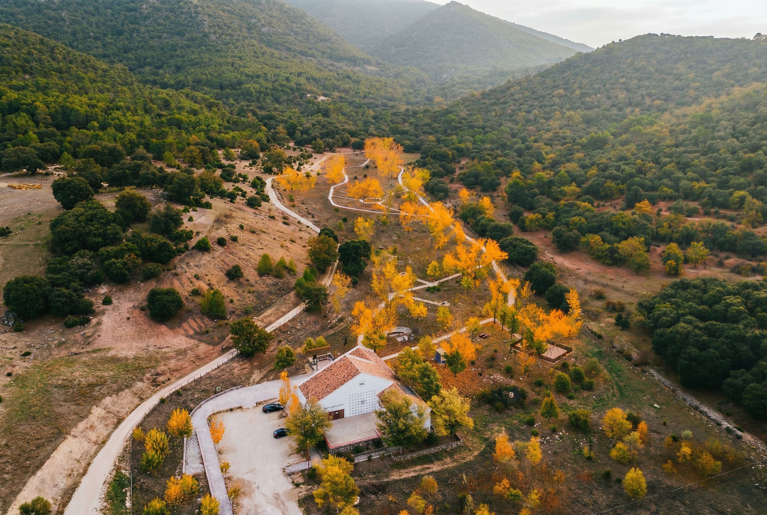 Vista aérea de un paisaje de montaña con una casa rural en primer plano, rodeada de caminos y árboles de tonos amarillos y anaranjados. El valle se extiende entre laderas cubiertas de vegetación, creando una escena amplia y tranquila.